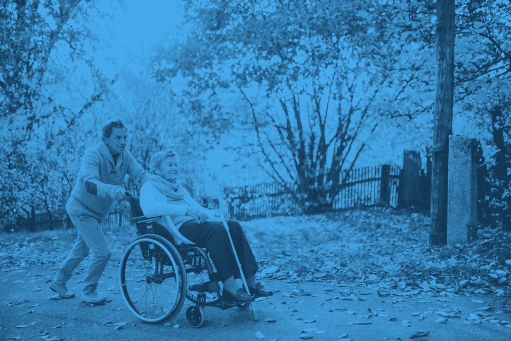 A guide for families pushing an elderly individual with dementia in a wheelchair outdoors on a path with autumn foliage.