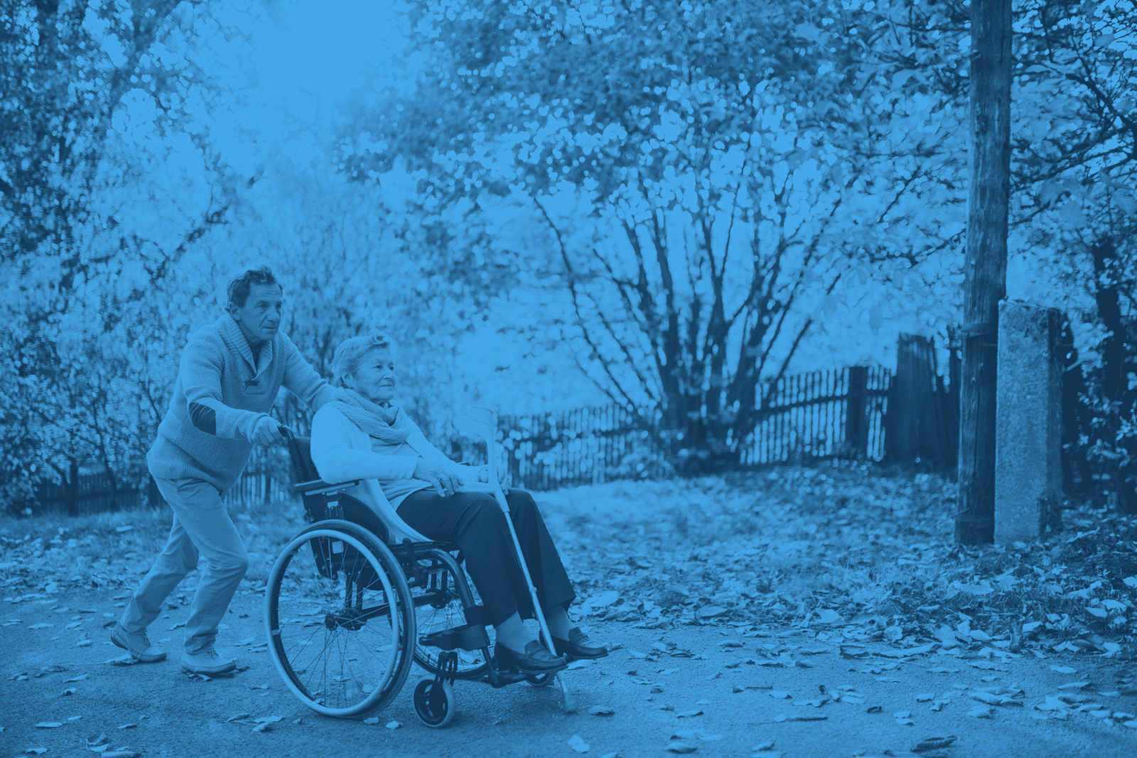 A guide for families pushing an elderly individual with dementia in a wheelchair outdoors on a path with autumn foliage.