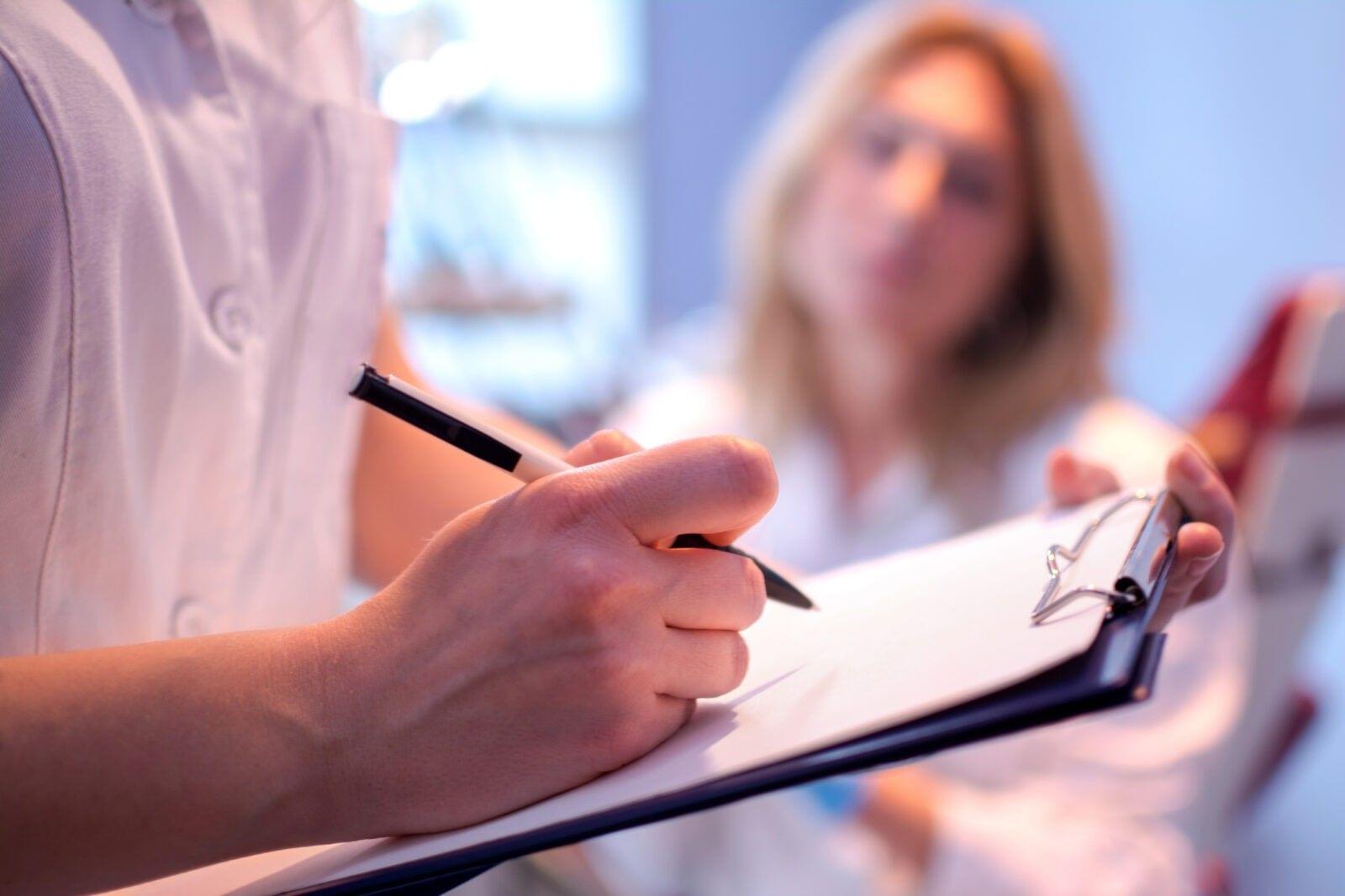 Health professional taking notes on a clipboard while consulting with a patient about new research.