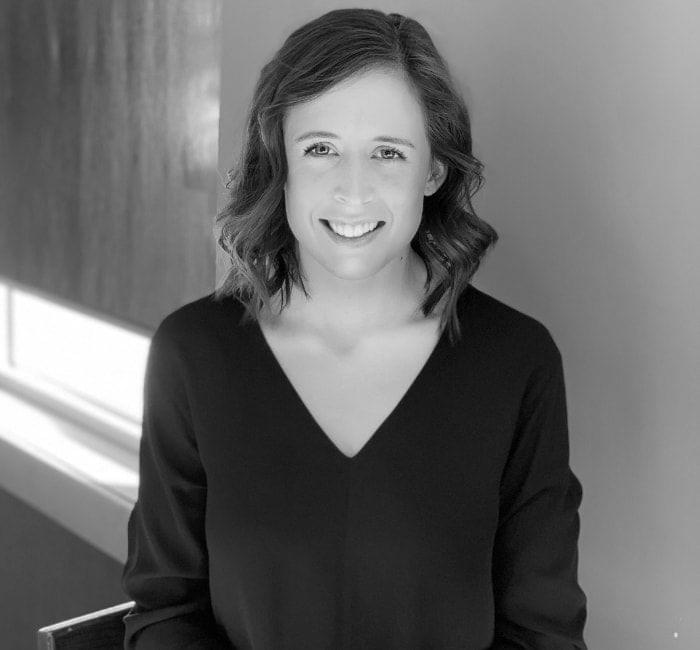 A black and white portrait of a smiling woman with shoulder-length hair, wearing a v-neck top.
