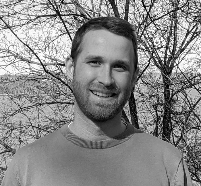 A man smiling in a black and white photo with trees and a water body in the background.