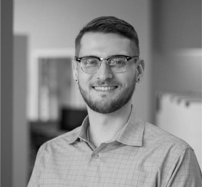 A smiling man wearing glasses and a checked shirt in a grayscale photograph.
