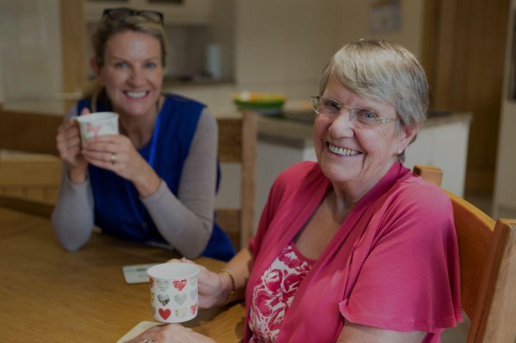 Two women smiling and enjoying a cup of tea at a kitchen table, integrating this simple pleasure into their daily workflow.