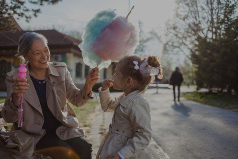 A joyful moment as a young girl, raised by her grandparent, offers a bite of her cotton candy to the elderly woman, both smiling during their outdoor encounter.