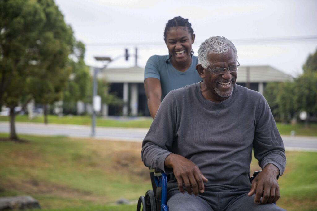 Woman smiling as she improves patient outcomes by pushing an elderly man in a wheelchair outdoors.
