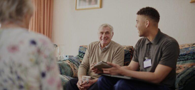Two individuals engaged in conversation about implementing family caregiver support while sitting in a living room, with one person holding a book and a cup, and the other holding a tablet.