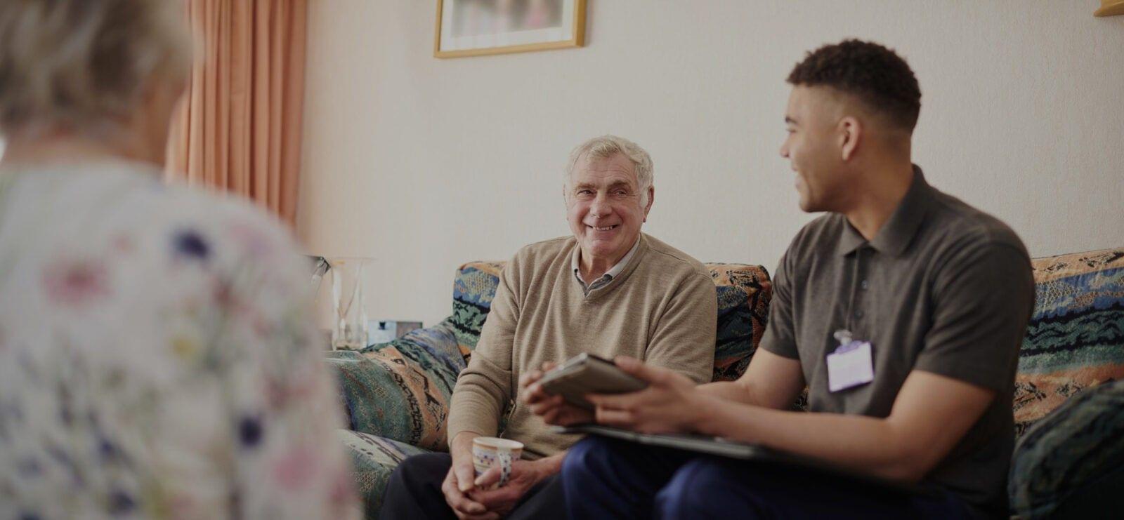 Two individuals engaged in conversation about implementing family caregiver support while sitting in a living room, with one person holding a book and a cup, and the other holding a tablet.