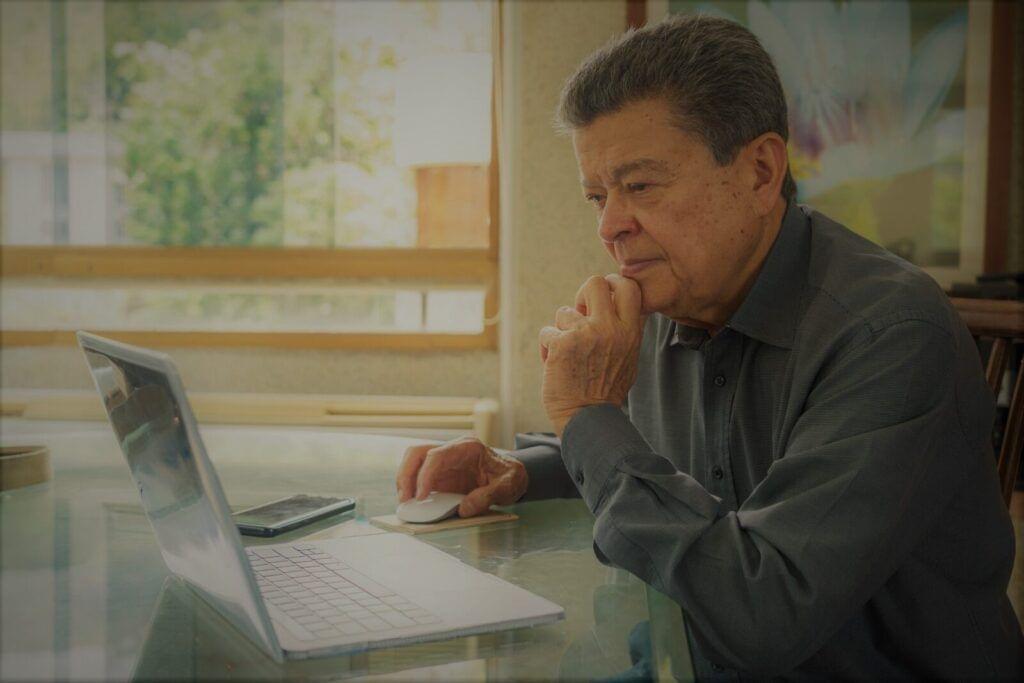 Senior man contemplating remote caregiver programs while using a laptop at a table.