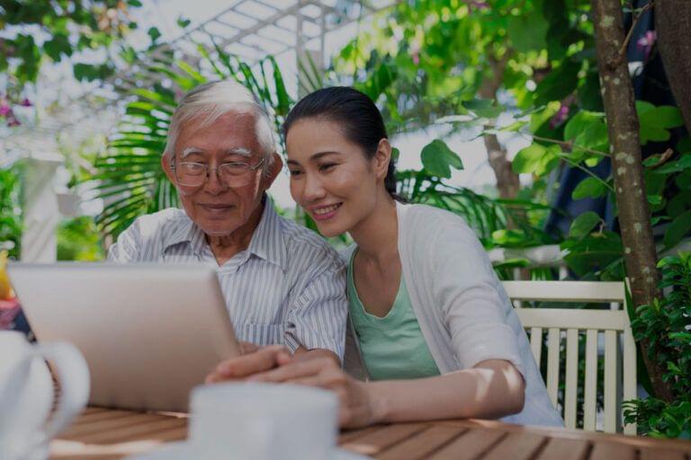 Senior man and young woman using a tablet together at an outdoor table to review the "Caregiving in the US 2015 vs 2020" comparison report.