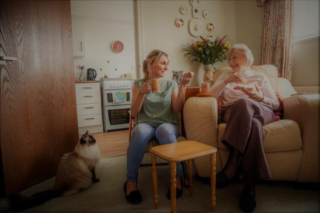 Two women enjoying a conversation with cups of tea in a cozy home setting with a cat nearby, and Alzheimer's Music Connect playing softly in the background.