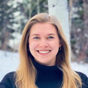 A woman with long blonde hair, representing various career opportunities, smiling at the camera, wearing a black turtleneck sweater, with a snowy background and trees partially visible behind her.