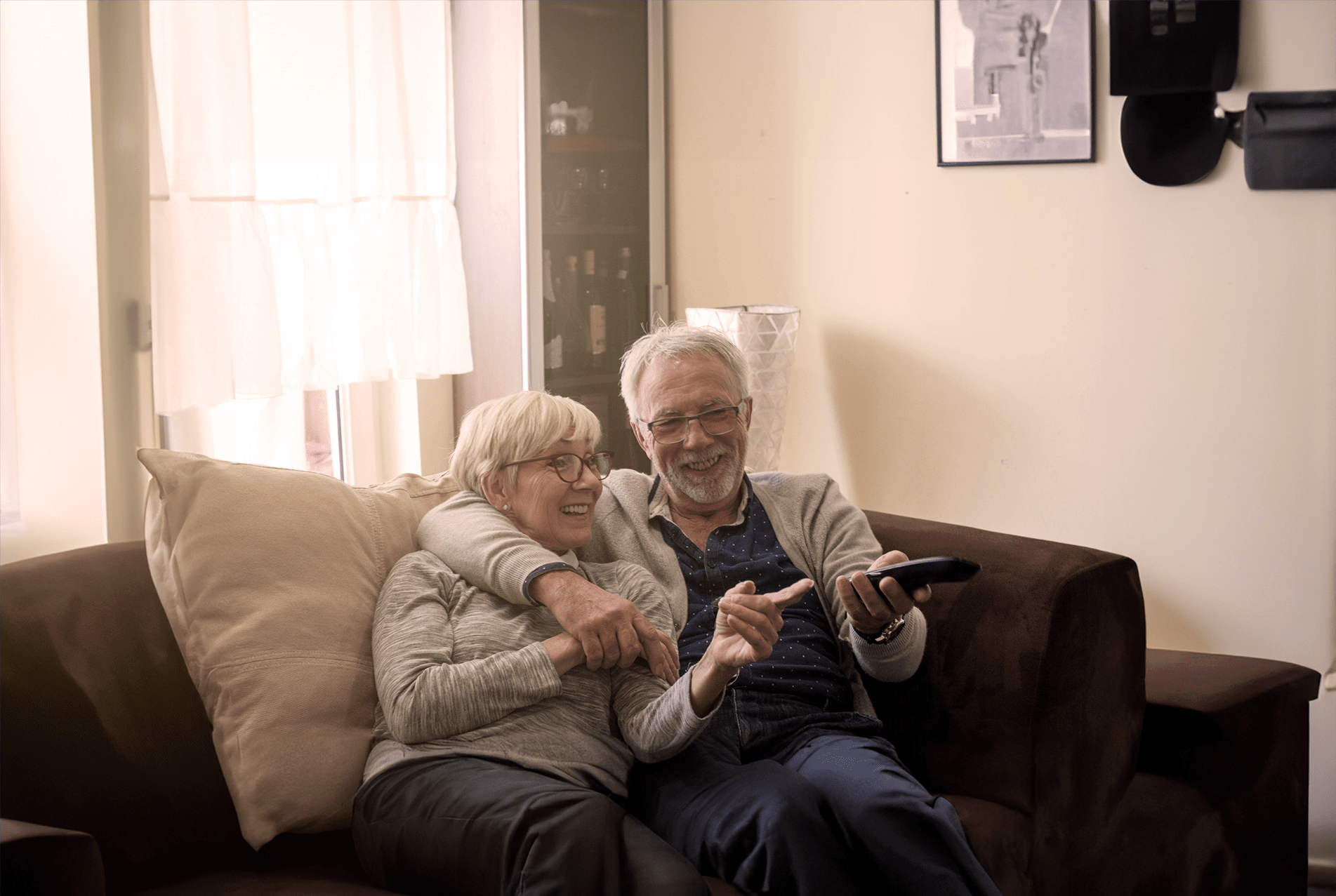 Elderly couple sharing a joyful moment on a couch, with the man holding a remote control, browsing through Trualta press releases.