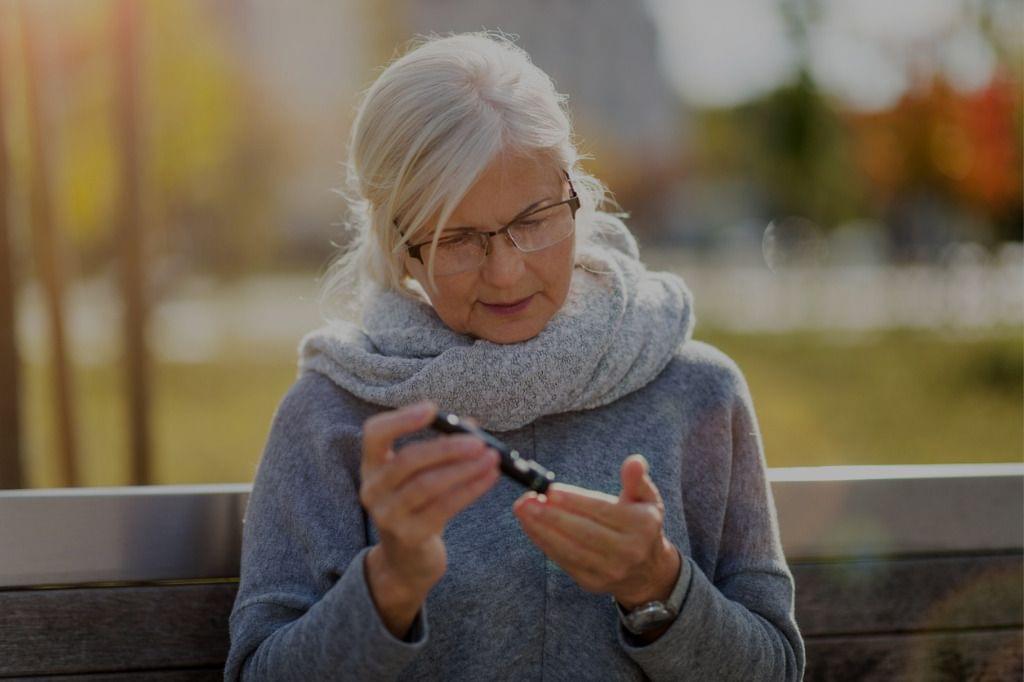 Senior woman checking blood sugar level with glucometer while sitting on a bench outdoors, managing her new diabetes.