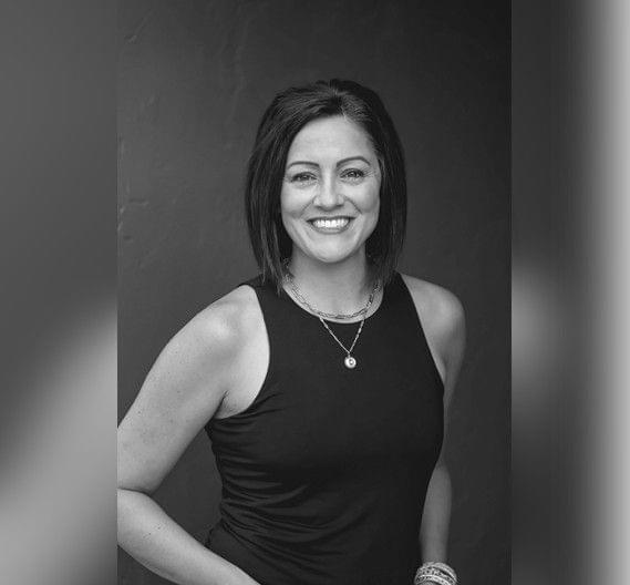 A smiling woman posing in a sleeveless black top against a gray background in a black-and-white photograph.
