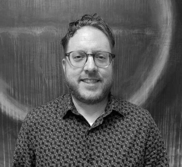 A man with glasses smiling in front of a textured background in a black and white photo.