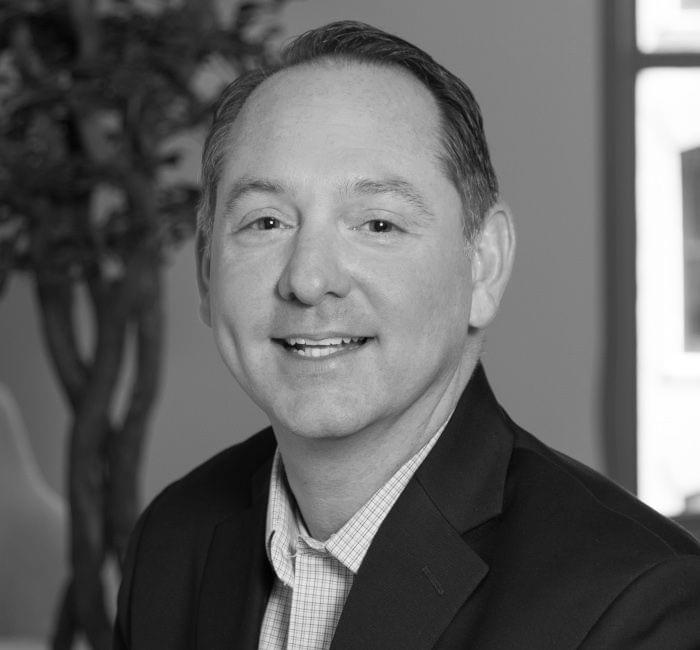 A black and white headshot of a smiling man in professional attire.