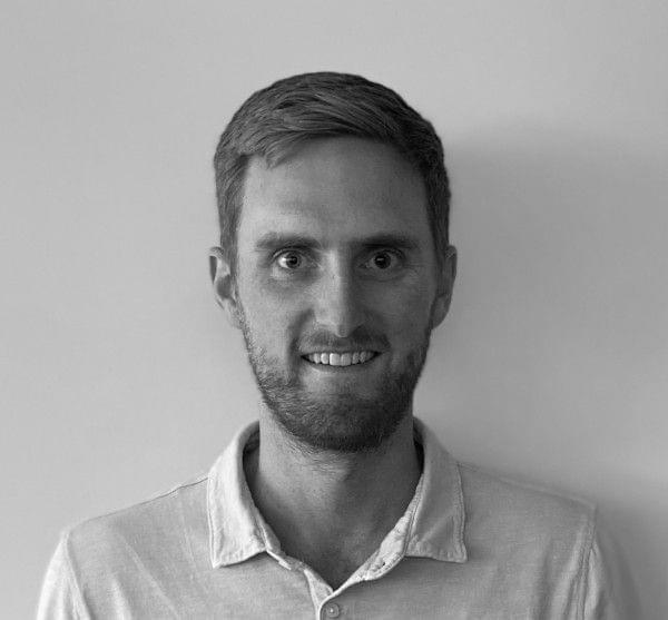 A black and white headshot of a man with a beard smiling at the camera.