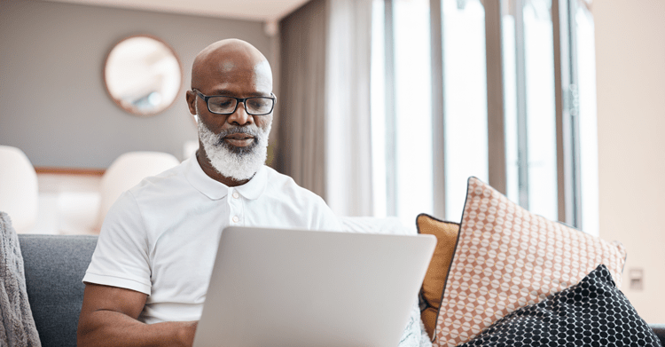 A man with glasses working on a laptop while sitting on a couch, using the Accessible Trualta Caregiver Portal.