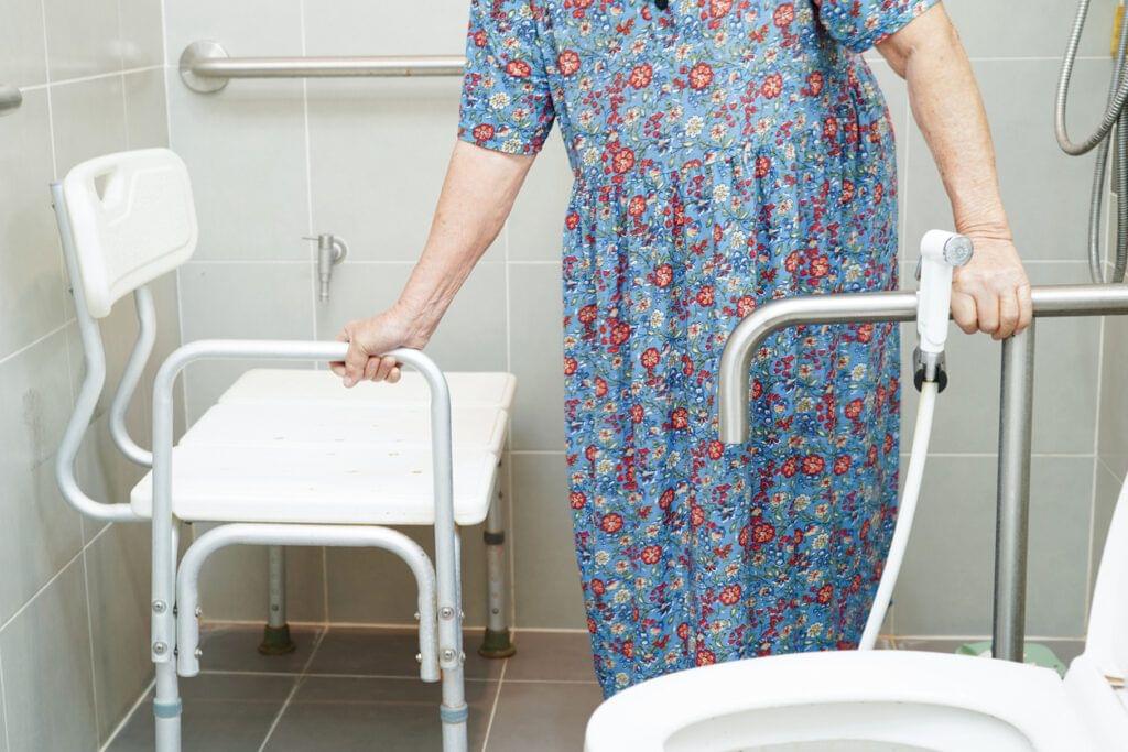 An older adult woman holds on to a grab bar with one hand and the shower chair arm rest with the other hand. Her face is not visible.