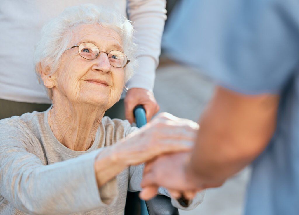 An older adult woman with glasses in a wheelchair holds the hands of a caregiver and looks up at them with a closed-lipped smile on her face.